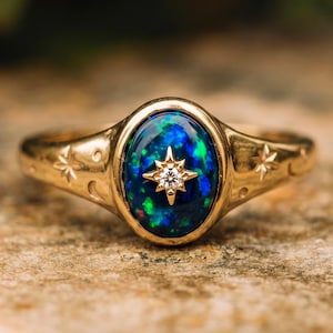 May include: A gold ring featuring an oval-shaped dark blue opal with a central star-shaped setting containing a small clear gemstone. The band is engraved with small star and crescent moon motifs on both sides of the central opal setting.
