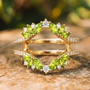 May include: A gold ring guard featuring two rows of marquise-cut peridot gemstones and small round white accent stones. The design includes a central open space framed by the green stones, with pavé-set white stones along the thin gold bands.