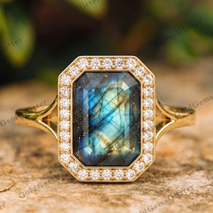 Op de afbeelding: Een gouden ring met een grote, rechthoekige, gefacetteerde labradoriet edelsteen met blauwe en groene iridiscentie. De middelste steen is omringd door een halo van kleine, ronde, helderwitte steentjes in een gouden zetting met een gespleten ringband.