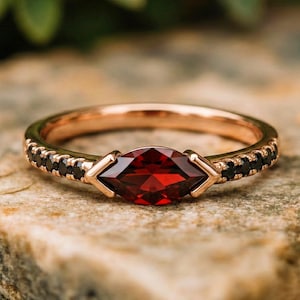 Puede incluir: Anillo de oro rosa con una piedra preciosa roja oscura con corte marquesa. El anillo tiene una banda con pequeñas piedras negras a cada lado de la piedra central. El anillo está sobre una superficie de piedra natural.