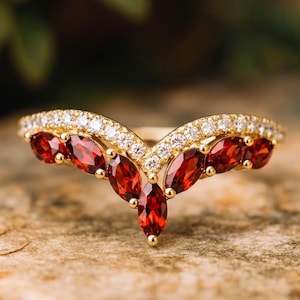 May include: A V-shaped gold ring featuring a row of marquise-cut deep red garnet gemstones set along the bottom edge, topped with a delicate band of small, clear, round-cut white stones that follow the chevron contour of the jewelry piece.