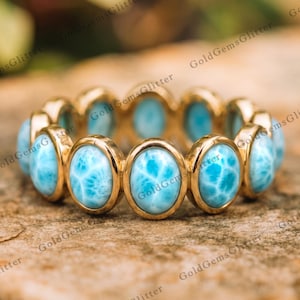 May include: A gold eternity band featuring a continuous row of oval-shaped, light blue Larimar gemstones. Each stone is set in a smooth gold bezel, creating a circular ring design with a polished finish and a vibrant, marbled blue appearance.