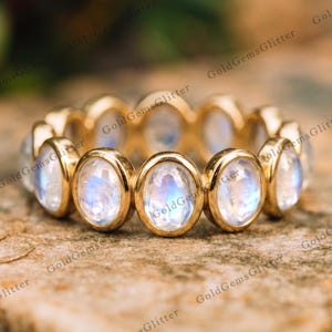 Puede incluir: Una alianza de oro con una hilera continua de piedras de luna ovaladas. Cada piedra de luna está engastada en un bisel de oro liso, mostrando un aspecto translúcido con una suave adularescencia azul. El anillo tiene un acabado en oro pulido.