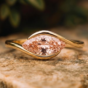 Pode incluir: Um anel de ouro com uma única pedra preciosa de cor pêssego em corte marquise, engastada horizontalmente. A banda de ouro lisa e polida curva-se em torno das extremidades pontiagudas da pedra, criando uma moldura que destaca a gema facetada.