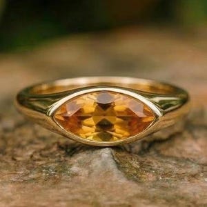 May include: A gold ring with an oval-shaped, yellow-orange gemstone. The ring is set on a rough, brown surface, with a blurred green background. The gemstone has multiple facets, reflecting light.