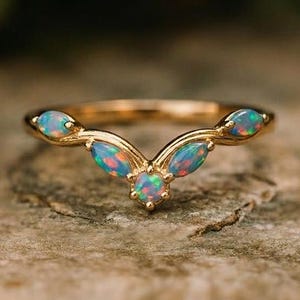 Puede incluir: Un anillo dorado con un diseño curvo, con cinco ópalos ovalados. Los ópalos muestran tonos azules, rojos y verdes. El anillo está sobre una superficie texturizada de color marrón claro.