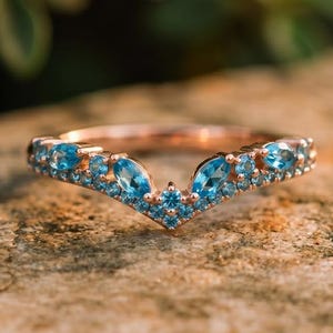 May include: A rose gold ring with a V-shaped design, set with numerous blue gemstones. The ring is displayed on a stone surface, with a blurred green backdrop.