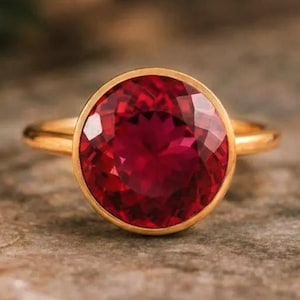 Puede incluir: Un anillo de oro con una gran piedra preciosa de rubí redonda y facetada. El rubí es de color rojo intenso y está engastado en un bisel de oro. El anillo está sobre una superficie texturizada de tonos neutros.