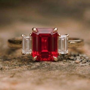 Puede incluir: Un anillo de oro con una gran gema rectangular de color rojo intenso, flanqueada por dos gemas claras rectangulares más pequeñas. El anillo está sobre una superficie marrón texturizada, mostrando el diseño y el contraste de color.