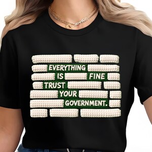 Puede incluir: Camiseta negra con el texto "EVERYTHING IS FINE TRUST YOUR GOVERNMENT" en blanco y con textura. El texto está sobre un fondo verde.