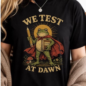 Op de afbeelding: Zwart T-shirt met een retro illustratie van een kikker met een cape, een potlood en een boek met het woord "EXAM". De tekst erboven luidt "WE TEST" en daaronder "AT DAWN."