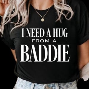 May include: Black t-shirt with the white text "I NEED A HUG FROM A BADDIE." The shirt has short sleeves and a crew neck. The model is wearing blue jeans and a gold necklace.
