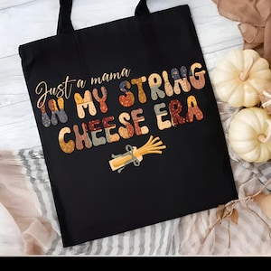 May include: Black tote bag featuring the phrase "Just a mama in my string cheese era" in patterned letters. A string cheese graphic is below the text. The bag has black handles and is on a light wood surface.