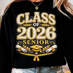 Op de afbeelding: Zwarte sweatshirt met de tekst "CLASS OF 2026 SENIOR" in goud- en crèmekleurige geborduurde letters. Het ontwerp bevat een afstudeerhoed, rollen en laurierbladeren, ter ere van de afstudeerklas van 2026.