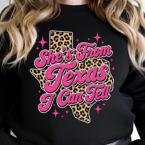 Puede incluir: Sudadera negra con un gráfico rosa y blanco que dice "She's From Texas I Can Tell". El gráfico presenta un contorno de Texas con estampado de leopardo y acentos de estrellas rosas. La sudadera es de estilo casual.