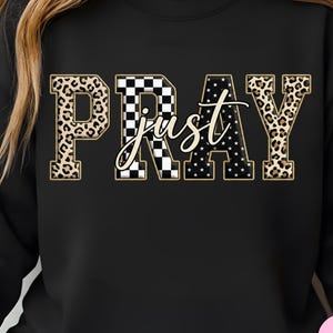 May include: Black sweatshirt with the word "PRAY" in a bold, decorative font. The letters have a leopard print, checkered, and star pattern. The word "just" is written in a cursive font over the word "PRAY".