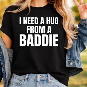 May include: Black t-shirt with the white text "I NEED A HUG FROM A BADDIE". The shirt has short sleeves and a crew neck. The model is wearing a denim jacket and blue jeans.