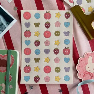 May include: A sheet of stickers with various designs, including buttons, strawberries, apples, hearts, butterflies, and stars. The stickers are in pastel colors and are arranged on a red and white striped background.