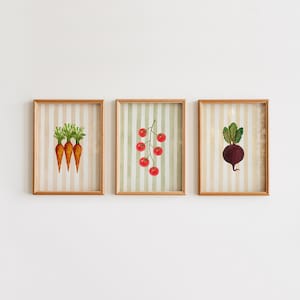 Könnte beinhalten: Drei gerahmte Aquarelldrucke von Gemüse. Einer zeigt Karotten, ein weiterer eine Traube Tomaten und der dritte eine Rote Bete. Jeder Druck hat einen gestreiften Hintergrund in neutralen Tönen. Die Rahmen sind hellholzfarben.