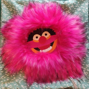 May include: A plush, pink, furry pillow featuring an embroidered cartoon face with a red nose, orange mouth, and white teeth. The pillow is set on a light green fabric with a white floral pattern.