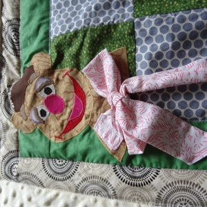 May include: A green and brown patchwork quilt square with a brown felt character with a pink nose and red mouth. The character has a pink bow with white swirls on it.