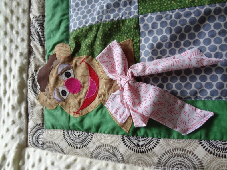 May include: A green and brown patchwork quilt square with a brown felt character with a pink nose and red mouth. The character has a pink bow with white swirls on it.