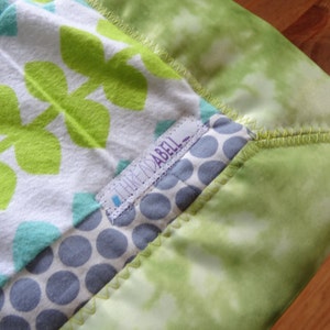 May include: A green, white, and blue baby blanket with a green leaf pattern and a label that says "Think Mabel".