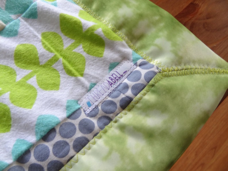 May include: A green, white, and blue baby blanket with a green leaf pattern and a label that says "Think Mabel".