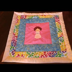 May include: A handmade quilt featuring a central pink square with an appliqued figure. The quilt has a blue patterned layer, a colorful floral border, and a white satin edge. The quilt is square-shaped.
