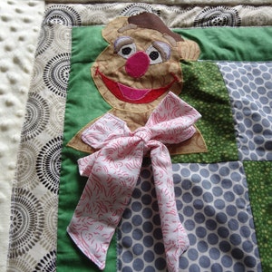 May include: A brown and tan felt puppet of a character with a pink nose and a pink bow tie. The puppet is on a patchwork quilt with green, brown, and gray fabric.