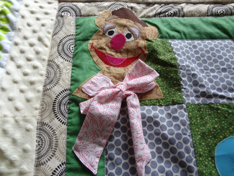 May include: A brown and tan felt puppet of a character with a pink nose and a pink bow tie. The puppet is on a patchwork quilt with green, brown, and gray fabric.