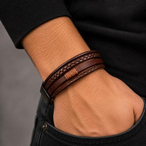 May include: A multi-strand bracelet featuring dark brown leather cords, including a braided strand and a smooth flat strand, secured with a black metal clasp and accented with a central copper-colored thread wrapping detail.