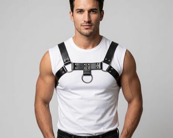Pode incluir: Um arnês de peito em couro preto com duas alças de ombro conectadas por uma faixa horizontal com rebites de metal prateado e três anéis de metal circulares, usado sobre uma camiseta branca sem mangas e calças jeans escuras.