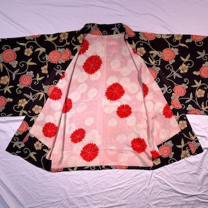 May include: A black kimono with a floral pattern featuring red and peach-coloured flowers and green leaves. The interior lining is light pink with large red and white floral designs. The kimono is spread out on a white surface.