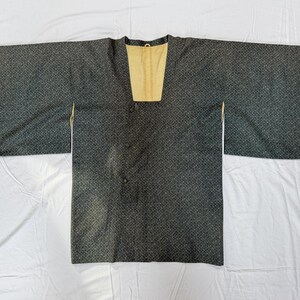 May include: A dark charcoal gray jacket with a fine, repeating geometric pattern across the fabric. The garment features wide, rectangular sleeves, a square neckline, and a light beige interior lining. Three small, round, dark fabric-covered buttons.