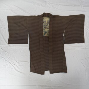 May include: A brown haori jacket featuring a subtle horizontal striped pattern across the fabric. The garment has wide sleeves and an open front design, revealing a contrasting inner lining with a detailed geometric and floral print pattern.