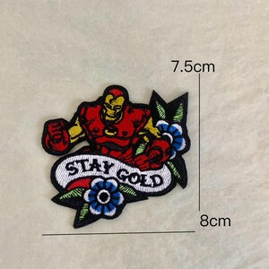 Op de afbeelding: Geborduurde patch met een rood en gele Iron Man figuur met de tekst "STAY GOLD" op een banner. De patch is ongeveer 8 cm breed en 7,5 cm hoog, met blauwe en groene bloemaccenten.