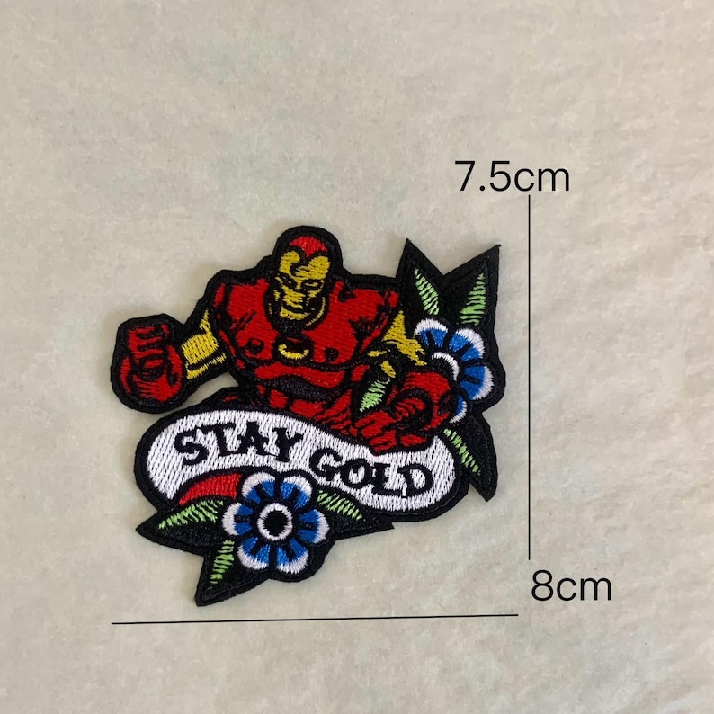 Op de afbeelding: Geborduurde patch met een rood en gele Iron Man figuur met de tekst "STAY GOLD" op een banner. De patch is ongeveer 8 cm breed en 7,5 cm hoog, met blauwe en groene bloemaccenten.