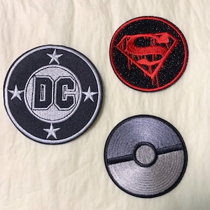 May include: Three circular embroidered patches. One features the DC Comics logo in black and white, another the Superman logo in red and black, and a third a grey and silver Pokeball design. Detailed stitching.