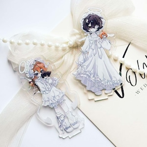 Peut inclure: Deux breloques en acrylique transparent représentant des personnages de style anime vêtus de blanc avec des ailes d'ange et des halos. Un personnage a les cheveux roux, l'autre a les cheveux noirs. Un rang de perles et un ruban blanc sont en arrière-plan.