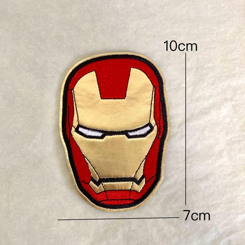 Op de afbeelding: Geborduurde patch met het Iron Man-masker in goud, rood en zwart. De patch is ongeveer 10 cm hoog en 7 cm breed. Het ontwerp toont de iconische helmvorm met witte oogdetails en een zwarte rand.