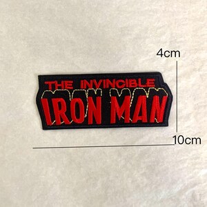 Op de afbeelding: Zwart en rood geborduurd embleem met de tekst "THE INVINCIBLE IRON MAN". De patch is rechthoekig en meet ongeveer 4 cm bij 10 cm. De tekst is rood met een gouden omtrek.