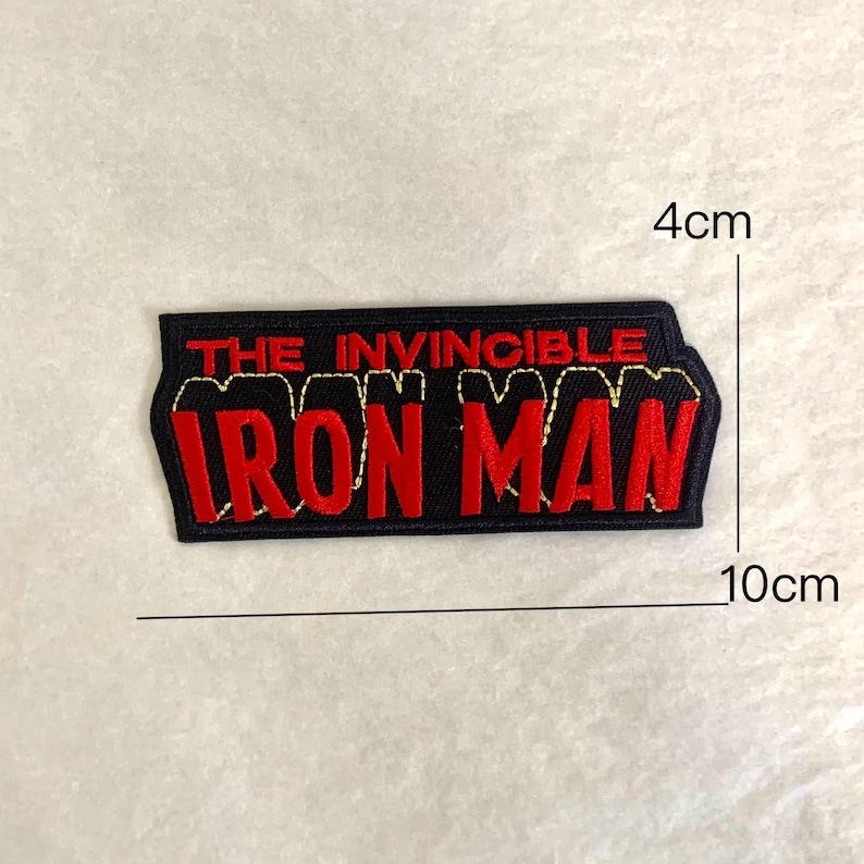 Op de afbeelding: Zwart en rood geborduurd embleem met de tekst "THE INVINCIBLE IRON MAN". De patch is rechthoekig en meet ongeveer 4 cm bij 10 cm. De tekst is rood met een gouden omtrek.