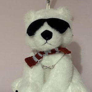 May include: A white plush polar bear keychain wearing black sunglasses and a red and white scarf. The bear has a black nose and paw pads, and a silver infinity symbol necklace. The keychain is attached to a silver keyring.