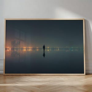 May include: A framed art print showing a person standing in a reflective body of water at dusk. The silhouette of the person is set against a backdrop of city lights and a dark, moody sky. The artwork is framed in a light-coloured wood.