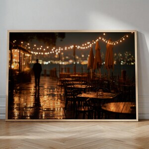 May include: A framed print of a nighttime outdoor restaurant scene. The image features a person walking on a wet wooden deck, with tables and chairs set up. Overhead, string lights illuminate the scene, with a city skyline in the background.