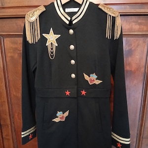 May include: Black military-style jacket with gold detailing, epaulets, and star and heart patches. The jacket has gold trim on the collar, cuffs, and front. Decorative gold buttons and red star accents complete the design. The jacket is hanging on a wooden hanger.