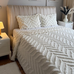 Peut inclure: Intérieur de chambre avec une tête de lit beige, une couverture de lit blanche à motif chevron texturé et des oreillers assortis. Trois impressions florales encadrées ornent le mur au-dessus du lit. Une petite table de chevet blanche avec une lampe et un tapis blanc moelleux.