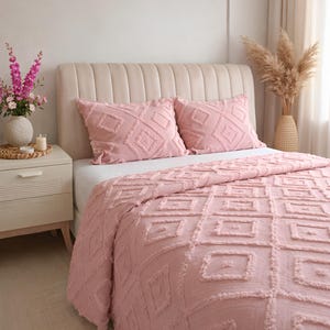 Peut inclure: Un couvre-lit rose et deux oreillers assortis avec un motif géométrique en losanges. La literie est sur un lit avec une tête de lit beige. Une table de chevet avec un vase de fleurs et une bougie est à gauche.