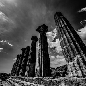 Può includere: Fotografia in bianco e nero di antiche colonne greche contro un cielo nuvoloso. Le colonne, alte, scanalate e usurate, presentano capitelli dettagliati. La prospettiva dal basso enfatizza l'altezza e la scala delle strutture.